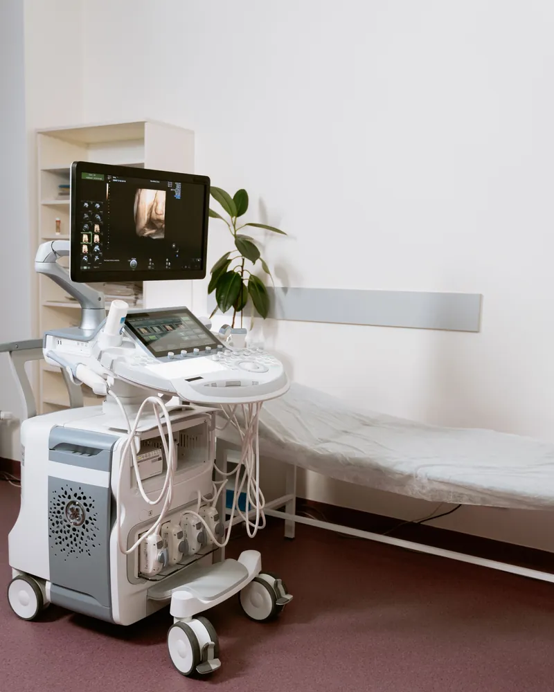 Aparelho de ultrassom com monitor ao lado da maca, numa sala de exame